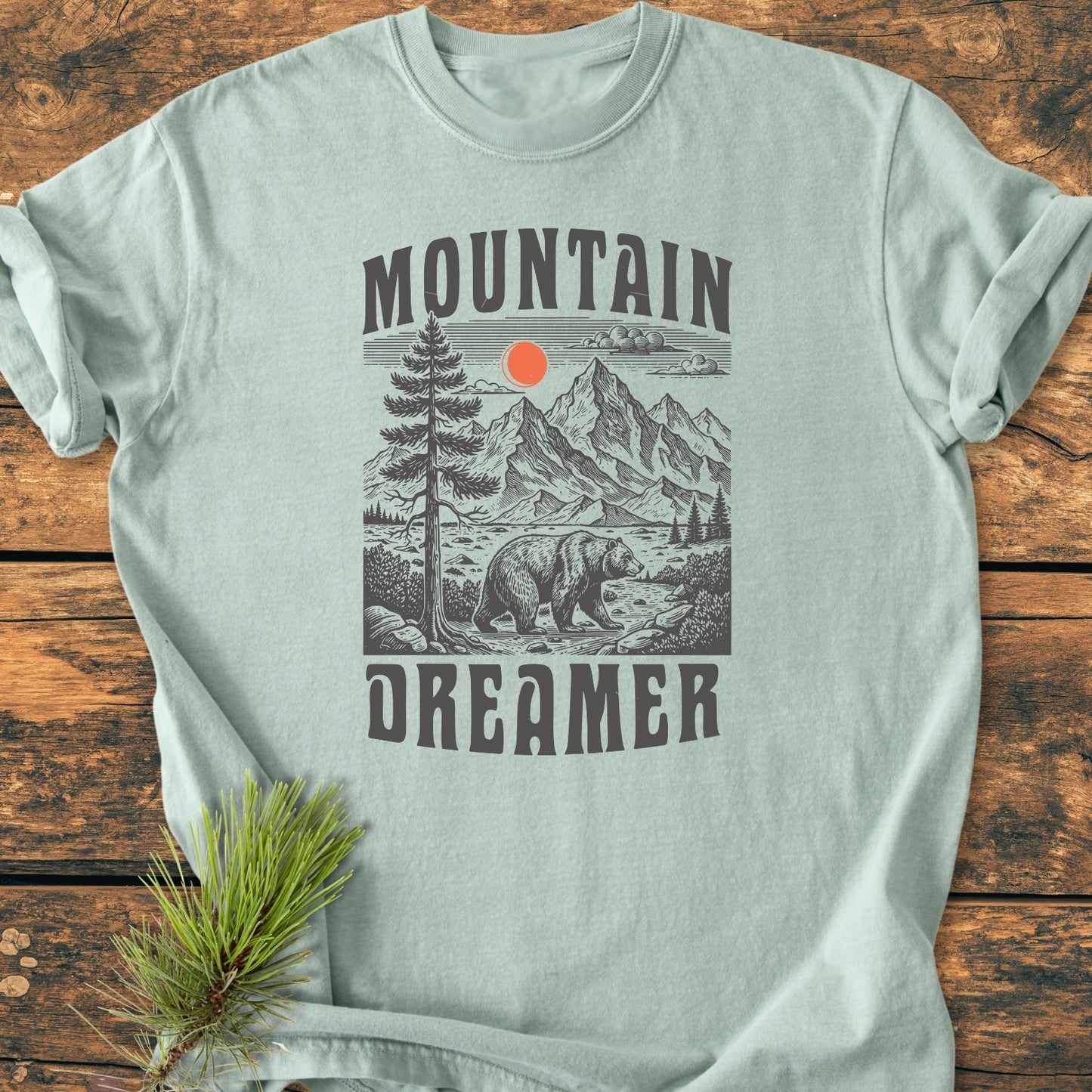 Mountain Dreamer - Vintage Wash Tee featuring a wood-cut outdoor scene with a bear, pine tree, mountains, and red sun. Laid on wooden surface with pine sprig styling.