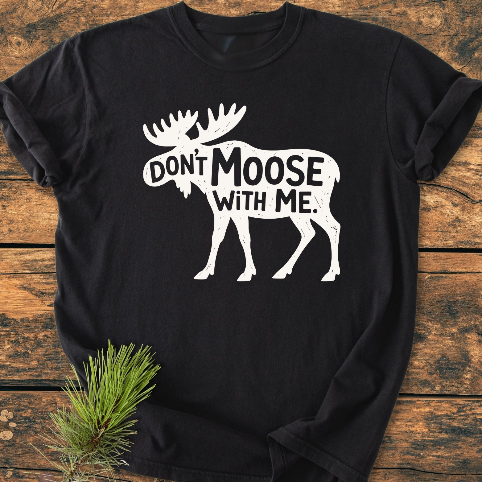 Don't Moose With Me - Vintage Wash Tee features a black shirt with a moose silhouette and punny text, laid on wood. Pine sprig accentuates the cozy, relaxed fit of this garment-dyed tee.