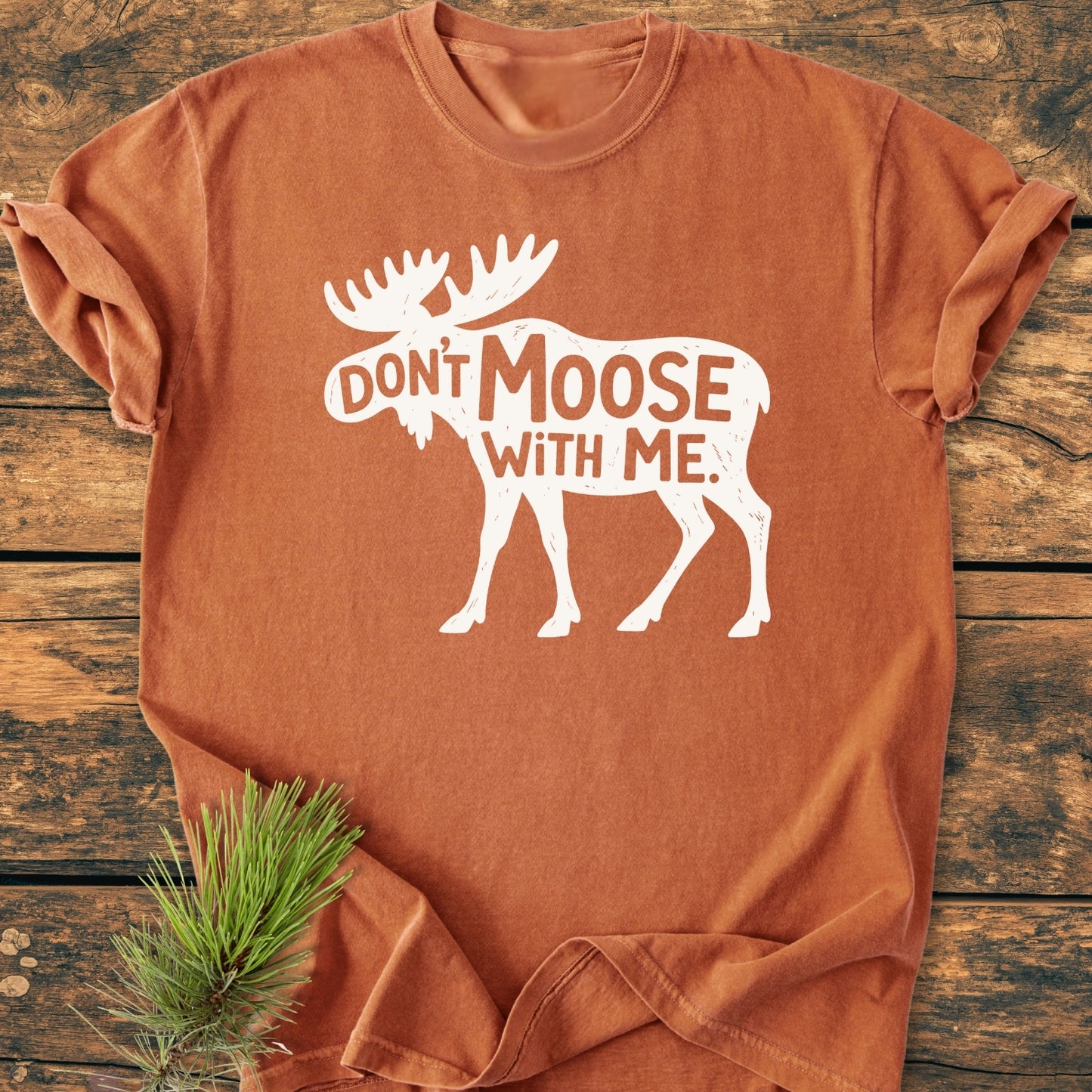Don't Moose With Me - Vintage Wash Tee in rust-orange, featuring a moose silhouette and slogan, laid flat on wood. Includes a pine branch accent, highlighting the tee's relaxed, durable design.