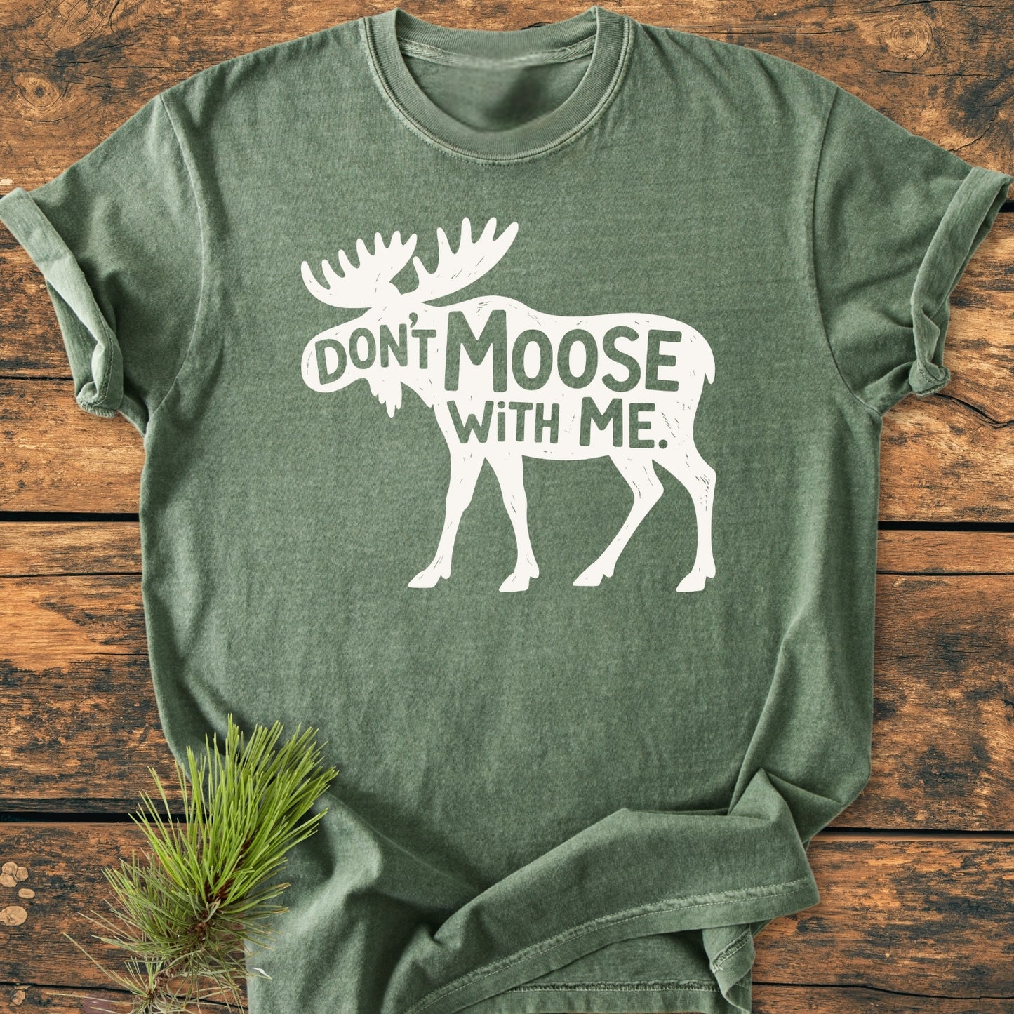 Don't Moose With Me - Vintage Wash Tee in heather-green with a white moose silhouette and playful text, laid on a wooden surface with a pine branch accenting the soft, relaxed fit design.