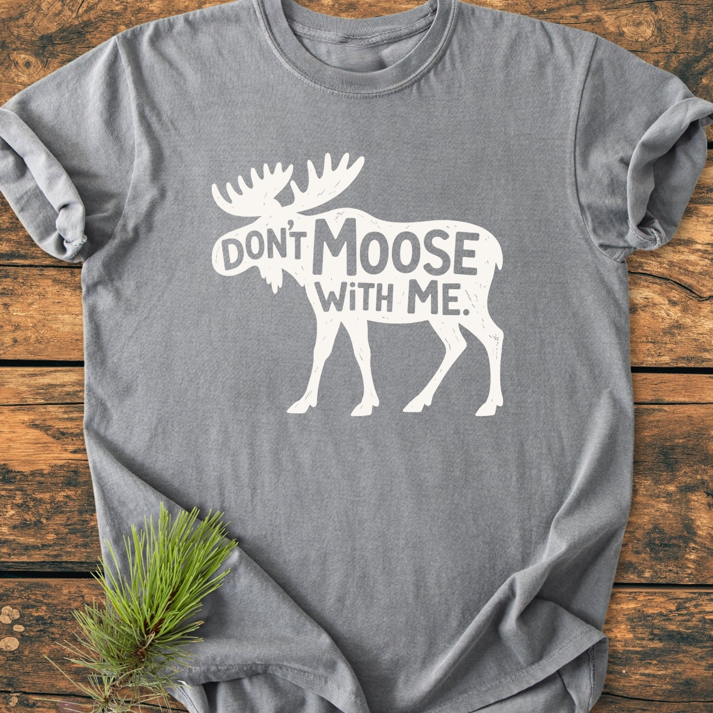 Heather-gray Don't Moose With Me - Vintage Wash Tee featuring a white moose silhouette with text, laid on wood with a pine sprig beside it. Crafted from soft, durable, garment-dyed cotton for comfort.