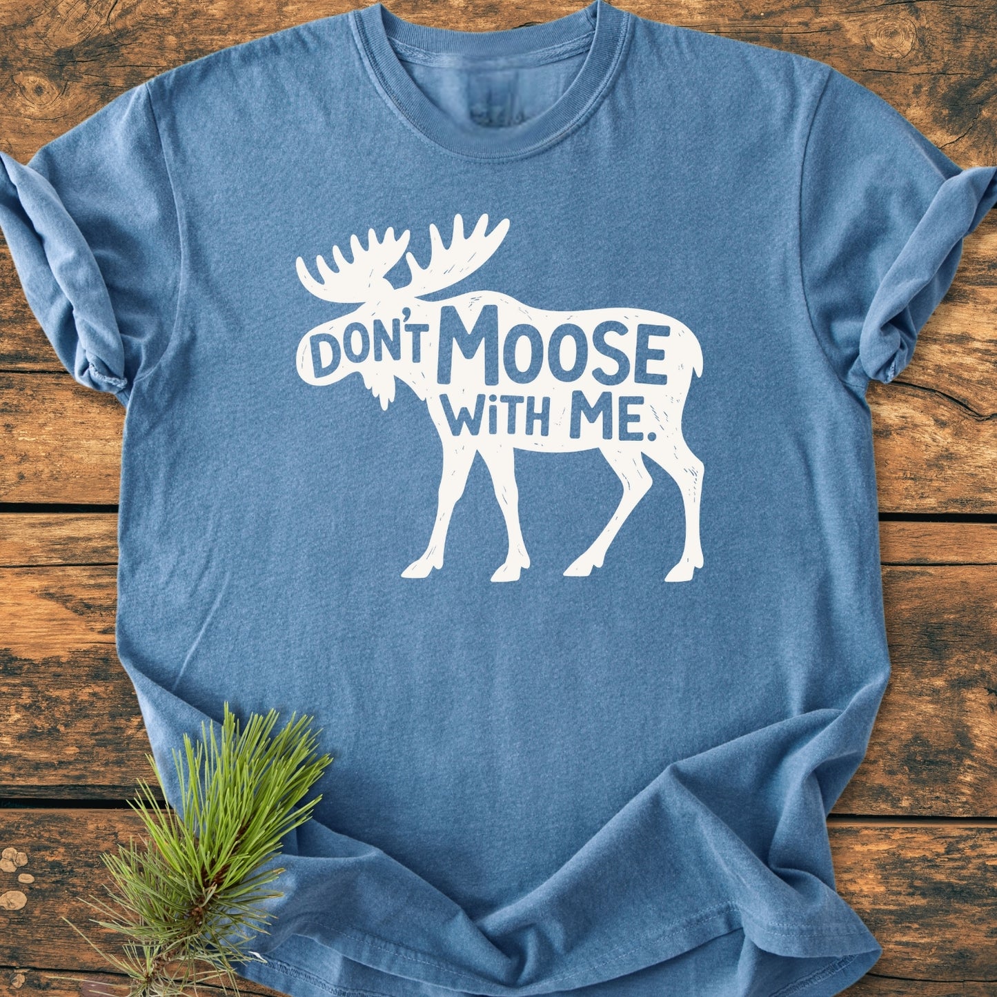 Don't Moose With Me - Vintage Wash Tee features a heather-blue design with a playful moose silhouette and slogan, laid on a wooden surface, accented by a rustic pine sprig.