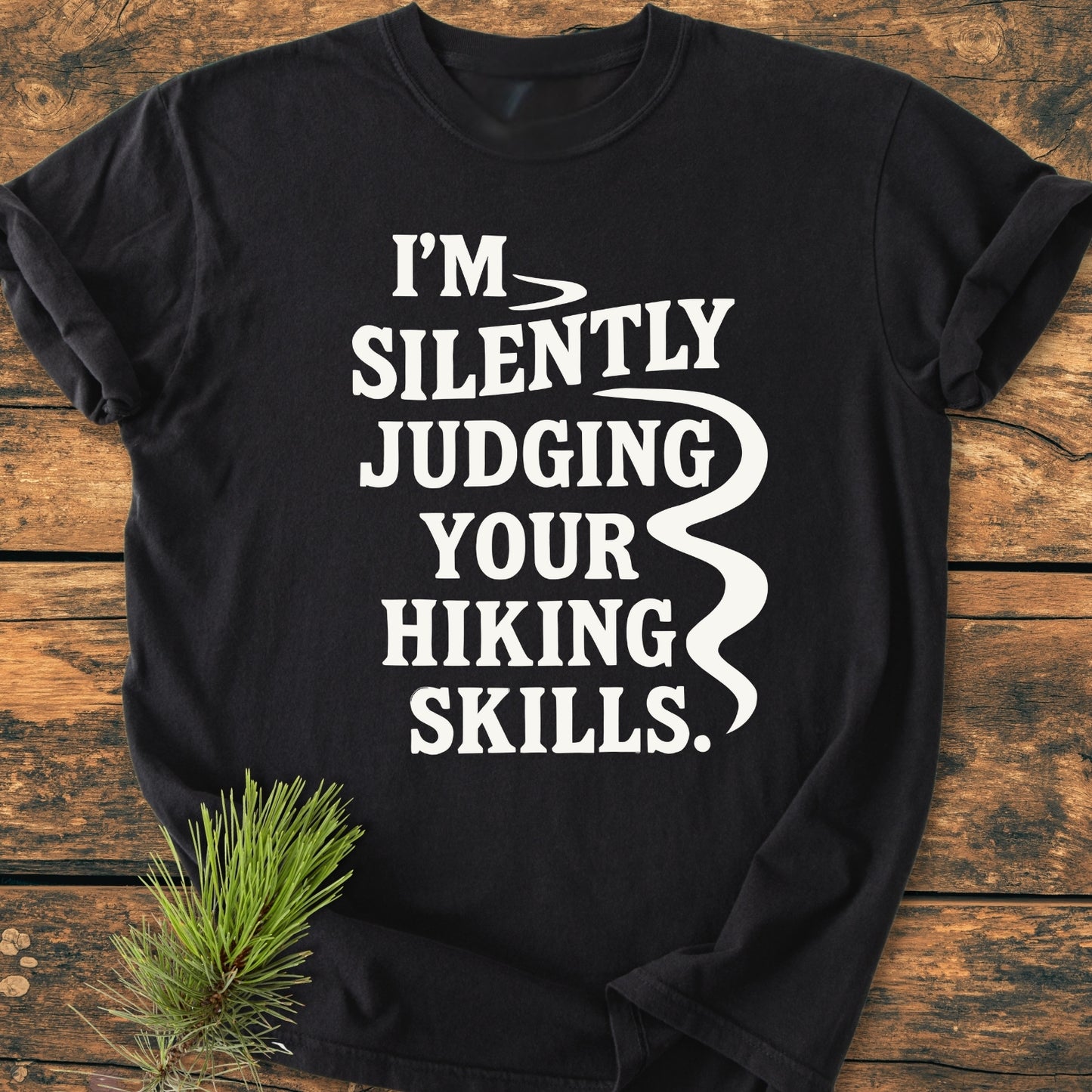 Silently Judging - Vintage Wash Tee features a black crew-neck design with bold white text and a stylized trail graphic, laid on wood with a pine sprig, showcasing its relaxed, durable, cotton fabric.