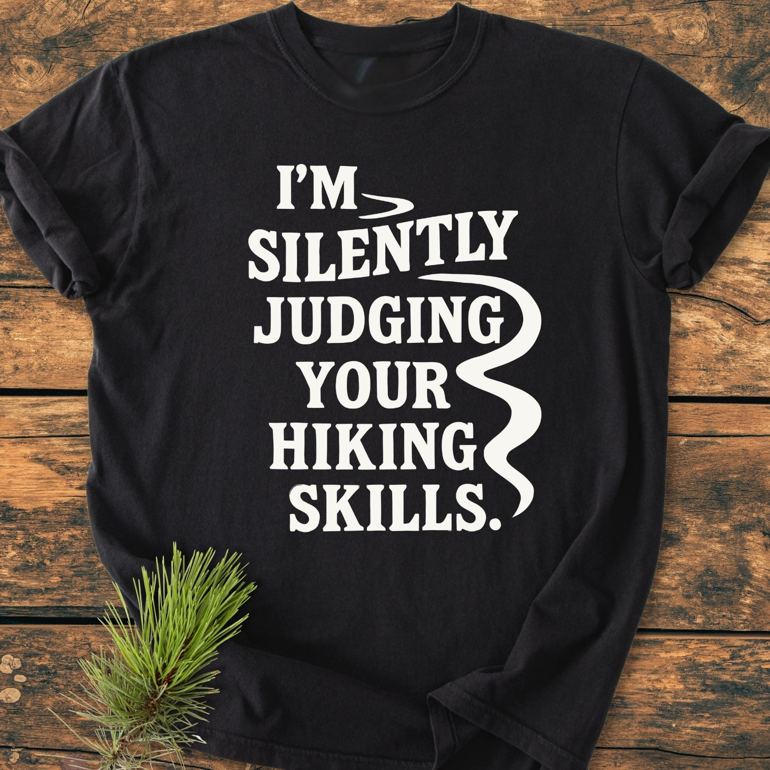 Silently Judging - Vintage Wash Tee features a black crew-neck design with bold white text and a stylized trail graphic, laid on wood with a pine sprig, showcasing its relaxed, durable, cotton fabric.
