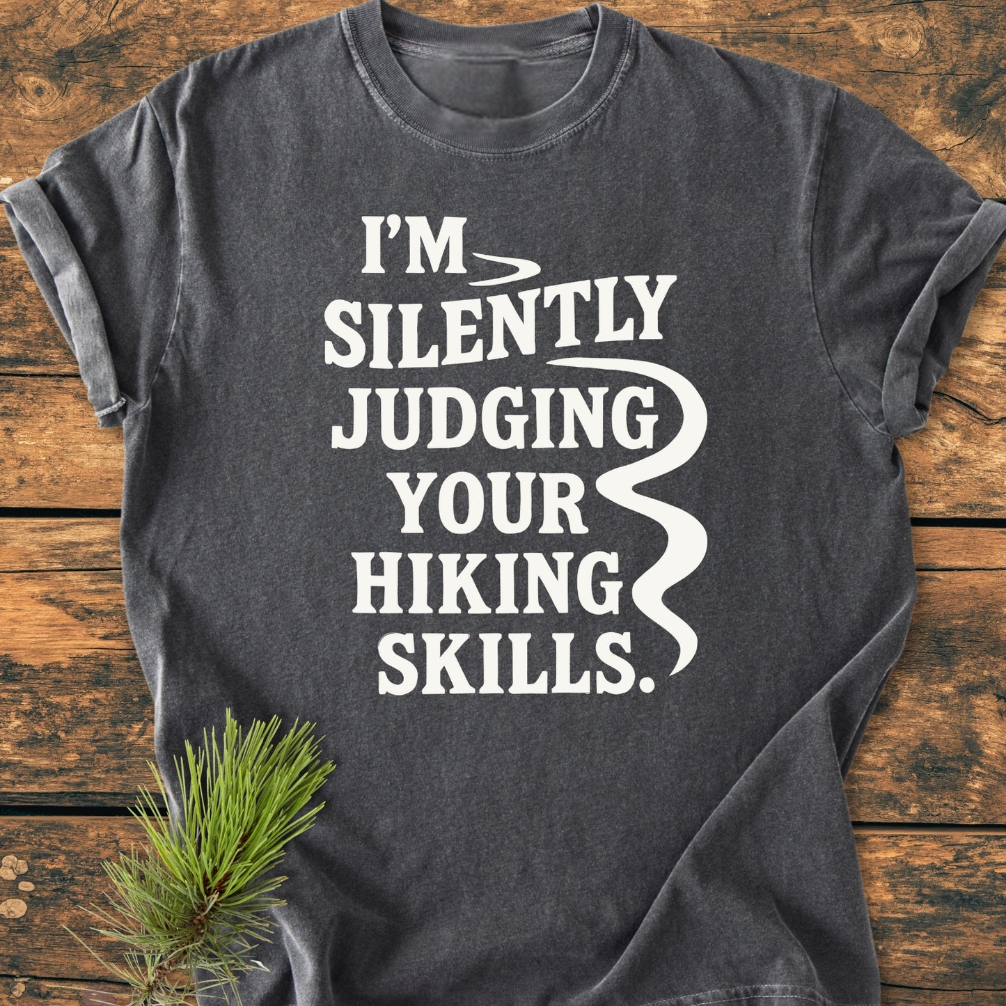 Silently Judging - Vintage Wash Tee featuring a bold white slogan on a charcoal-gray T-shirt, laid flat with a rustic pine sprig adjacent, highlighting its relaxed fit and durable, soft-washed fabric.