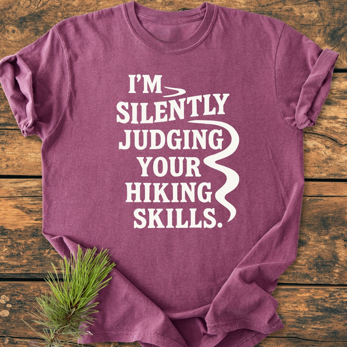 Silently Judging - Vintage Wash Tee in maroon, displaying bold white text I’M SILENTLY JUDGING YOUR HIKING SKILLS, folded on a wooden surface with an evergreen sprig beside it.