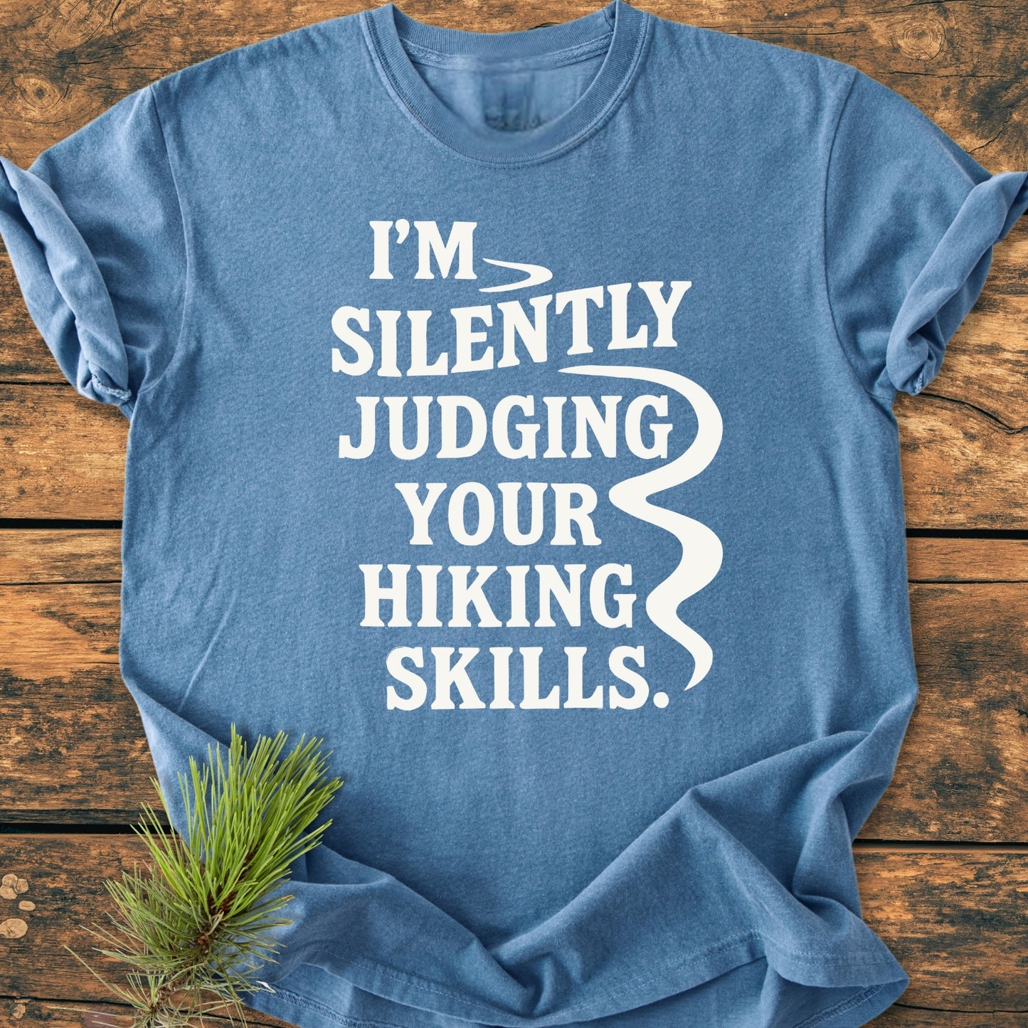 Silently Judging - Vintage Wash Tee in heather-blue on wood surface, features bold white text I’M SILENTLY JUDGING YOUR HIKING SKILLS, accented with a pine sprig for outdoorsy flair.