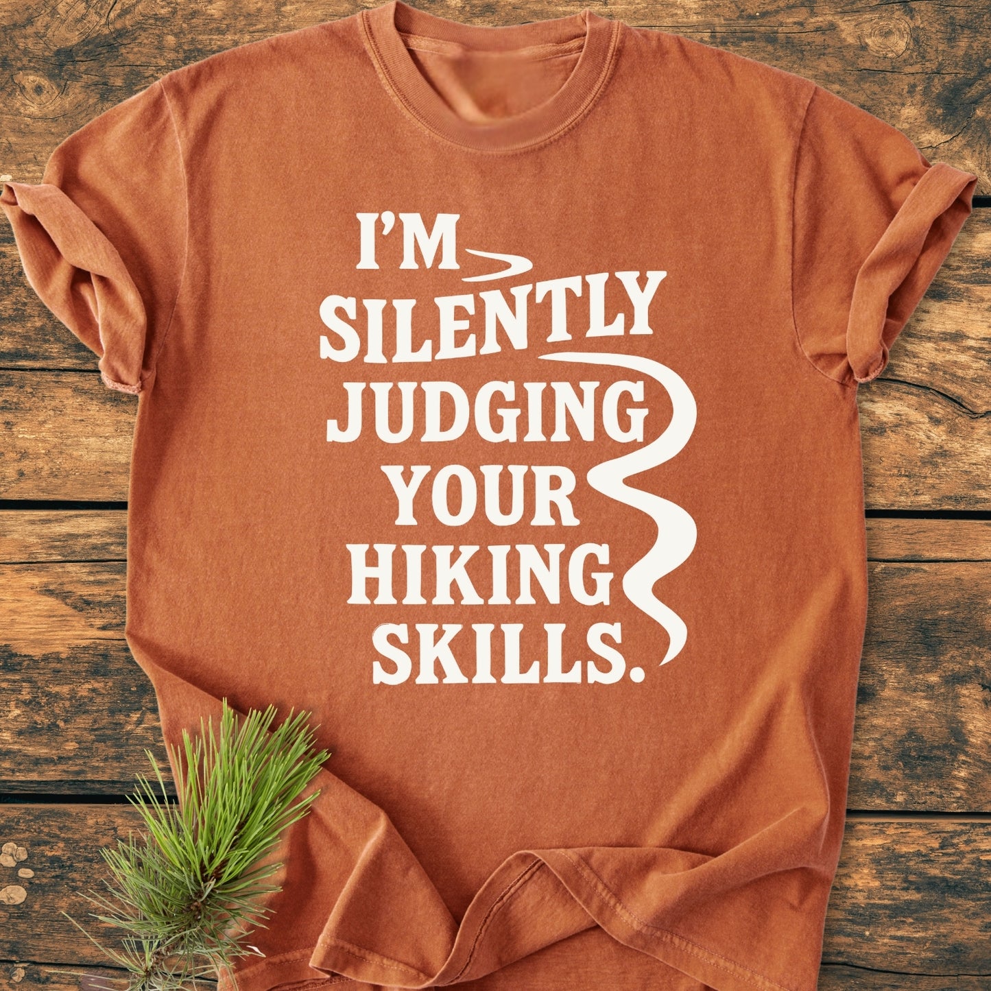 Silently Judging - Vintage Wash Tee laid on wood, features bold white text about hiking skills, complemented by a wavy line and pine sprig, emphasizing its outdoor theme and relaxed, cozy fit.