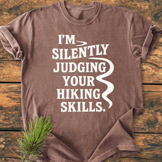 Silently Judging - Vintage Wash Tee on wooden surface, features bold white slogan with stylized swoosh. A pine twig rests on the shirt, emphasizing its relaxed, outdoor-inspired aesthetic.