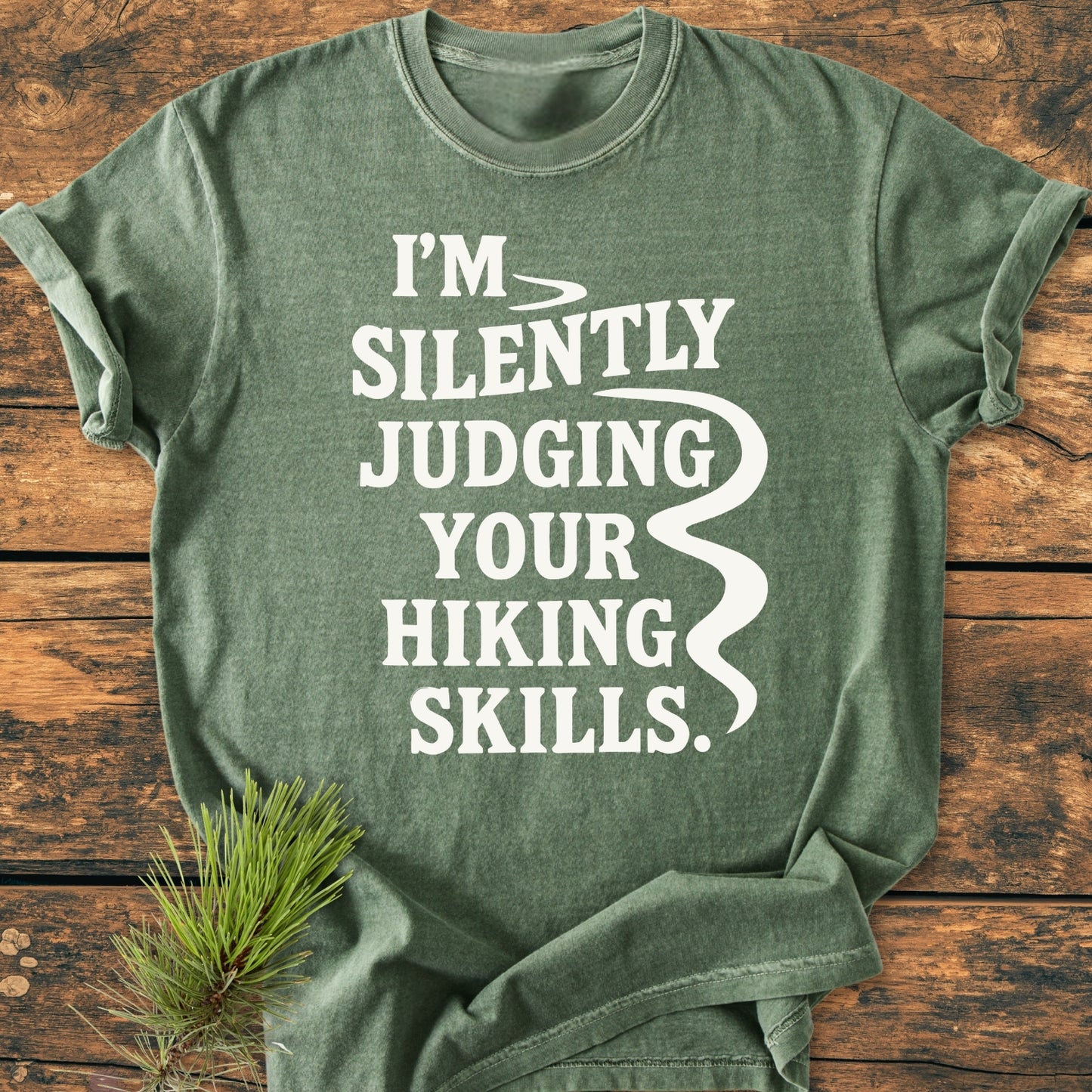 Silently Judging - Vintage Wash Tee, a forest-green crew-neck with rolled sleeves, features bold white text on a rustic wooden surface, accompanied by a pine sprig, enhancing its outdoorsy appeal.