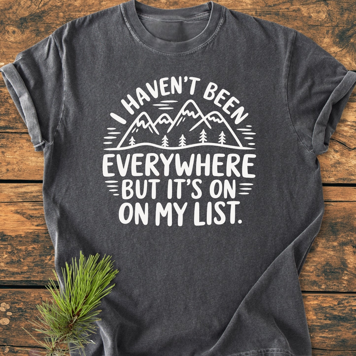 On My List - Vintage Wash Tee: Charcoal-gray with a white, hand-lettered mountain design, displayed on a wooden surface. Rolled sleeves and pine sprig enhance the outdoorsy, travel-inspired look.