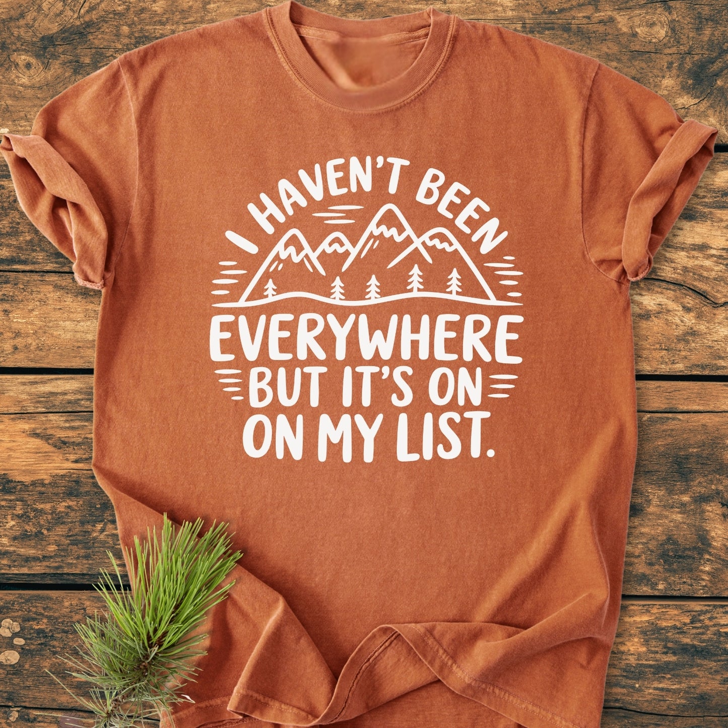 On My List - Vintage Wash Tee features a rust-orange, crew-neck design with a mountain-and-pine-tree graphic and travel slogan, laid flat with a pine sprig accentuating its outdoorsy charm.