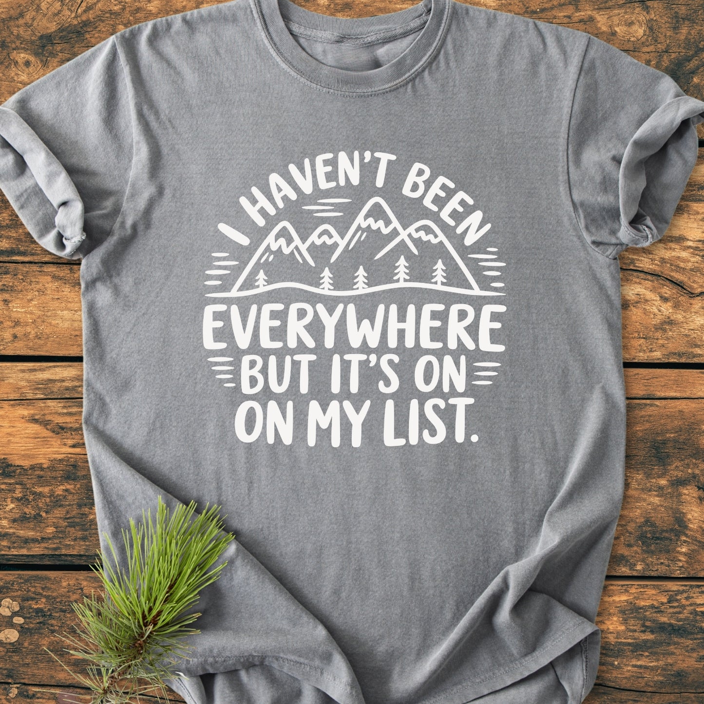 On My List - Vintage Wash Tee displayed on a wooden surface, featuring a white mountain scene and quote. A sprig of pine needles accents the heather-gray, relaxed-fit, garment-dyed T-shirt.