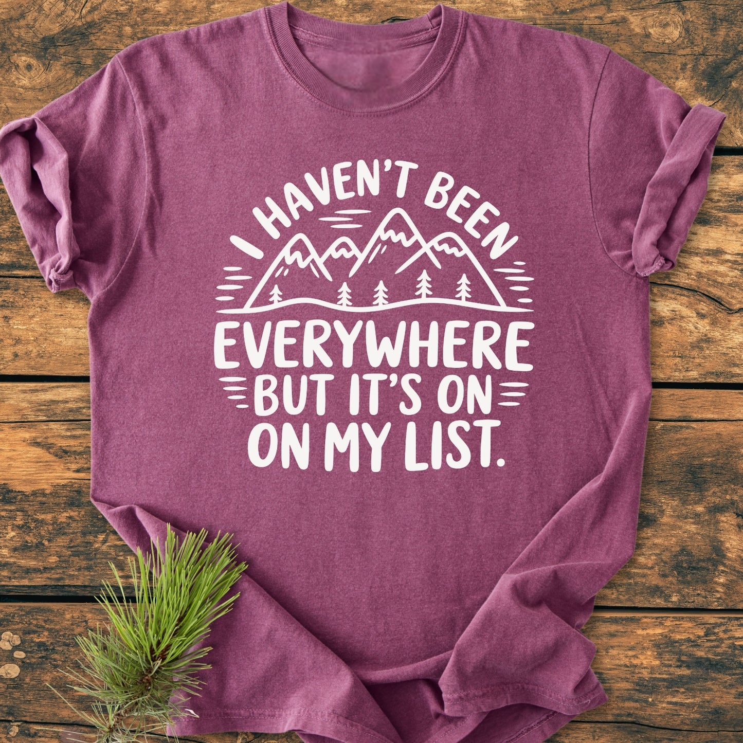 On rustic wood, a heathered purple On My List - Vintage Wash Tee with a mountain-and-pine-tree graphic and travel slogan, showcasing its relaxed fit and durable, soft-washed cotton fabric.