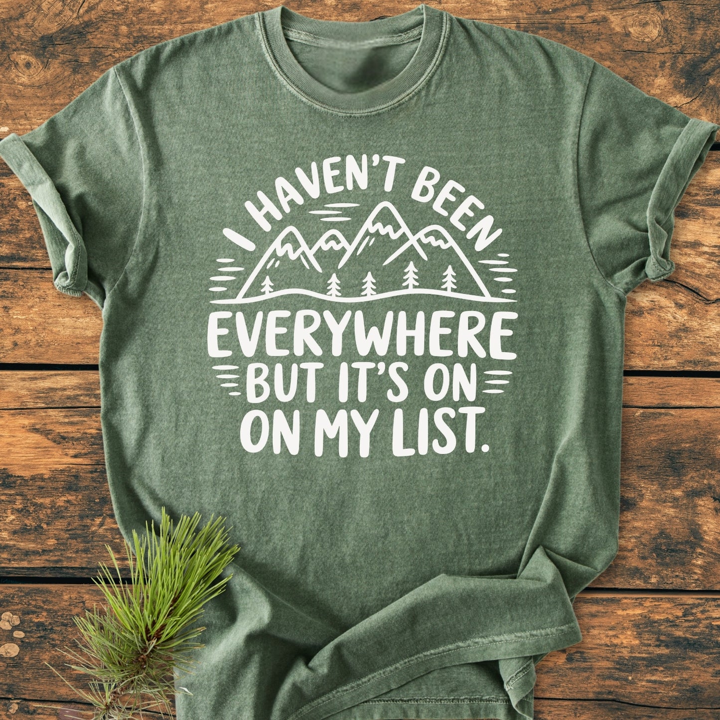 On My List - Vintage Wash Tee in heather-green, featuring a white mountain-and-tree design and phrase, styled with a pine branch, showcasing its relaxed fit and garment-dyed softness.
