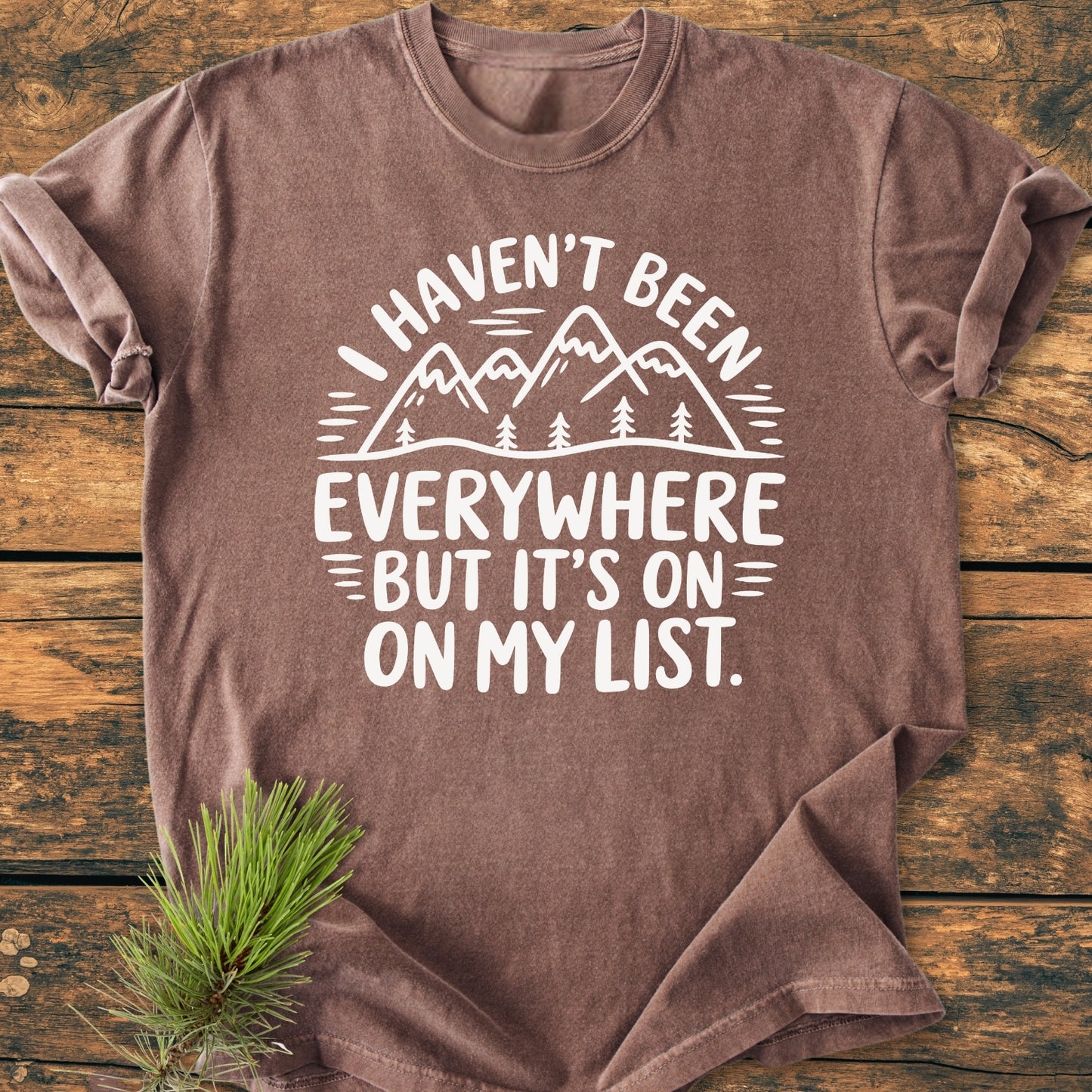 On My List - Vintage Wash Tee features a stylized mountain-and-trees graphic with the phrase, “I haven’t been everywhere but it’s on my list,” on a brown, short-sleeved shirt laid flat.