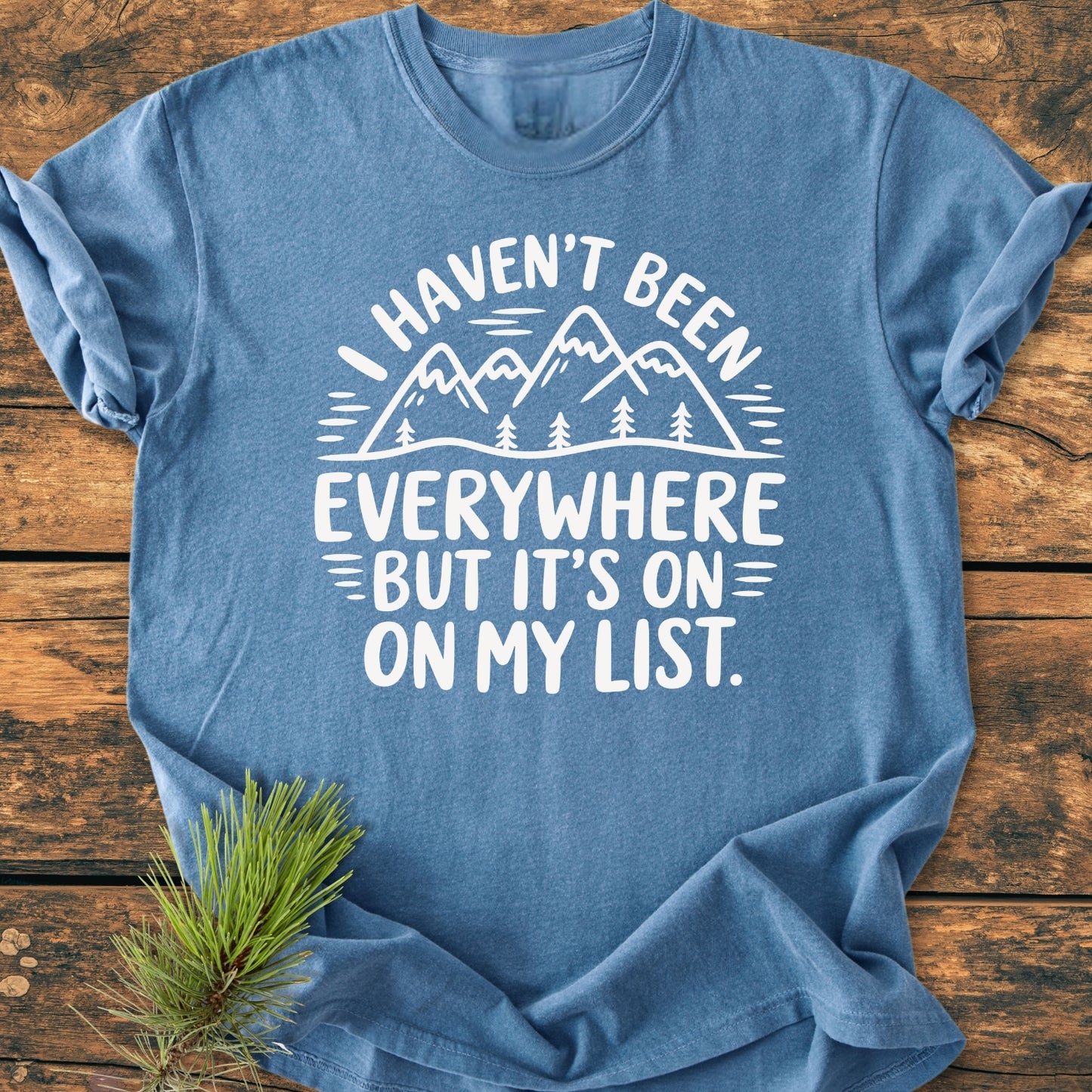 On My List - Vintage Wash Tee features a heather-blue color with a white mountain-and-tree graphic and slogan. Laid flat on wood, styled with a pine sprig under the hem.