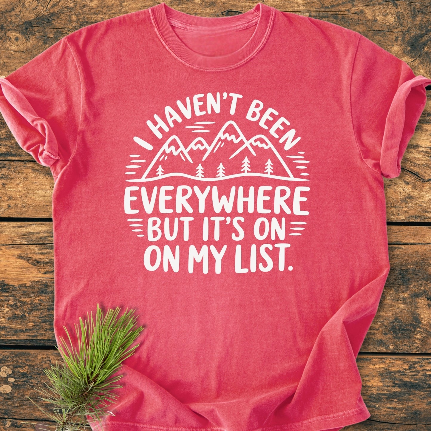 On My List - Vintage Wash Tee featuring a white graphic of mountain peaks and pine trees, with slogan text, laid on a rustic surface, highlighting its relaxed fit and garment-dyed fabric.
