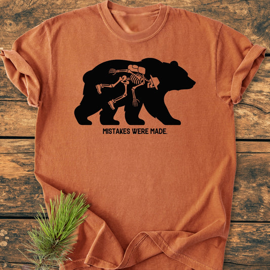 Mistakes Were Made - Vintage Wash Tee, featuring a bear with a skeleton hiker print, displayed on wood with a pine sprig. Made of soft, garment-dyed, ring-spun cotton for relaxed comfort.