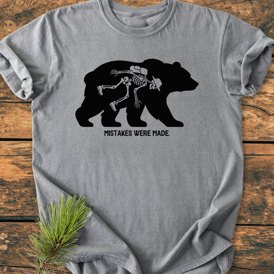 Mistakes Were Made - Vintage Wash Tee featuring a black bear silhouette with a human skeleton in hiking gear inside, displayed on a rustic surface with a pine sprig beside it.