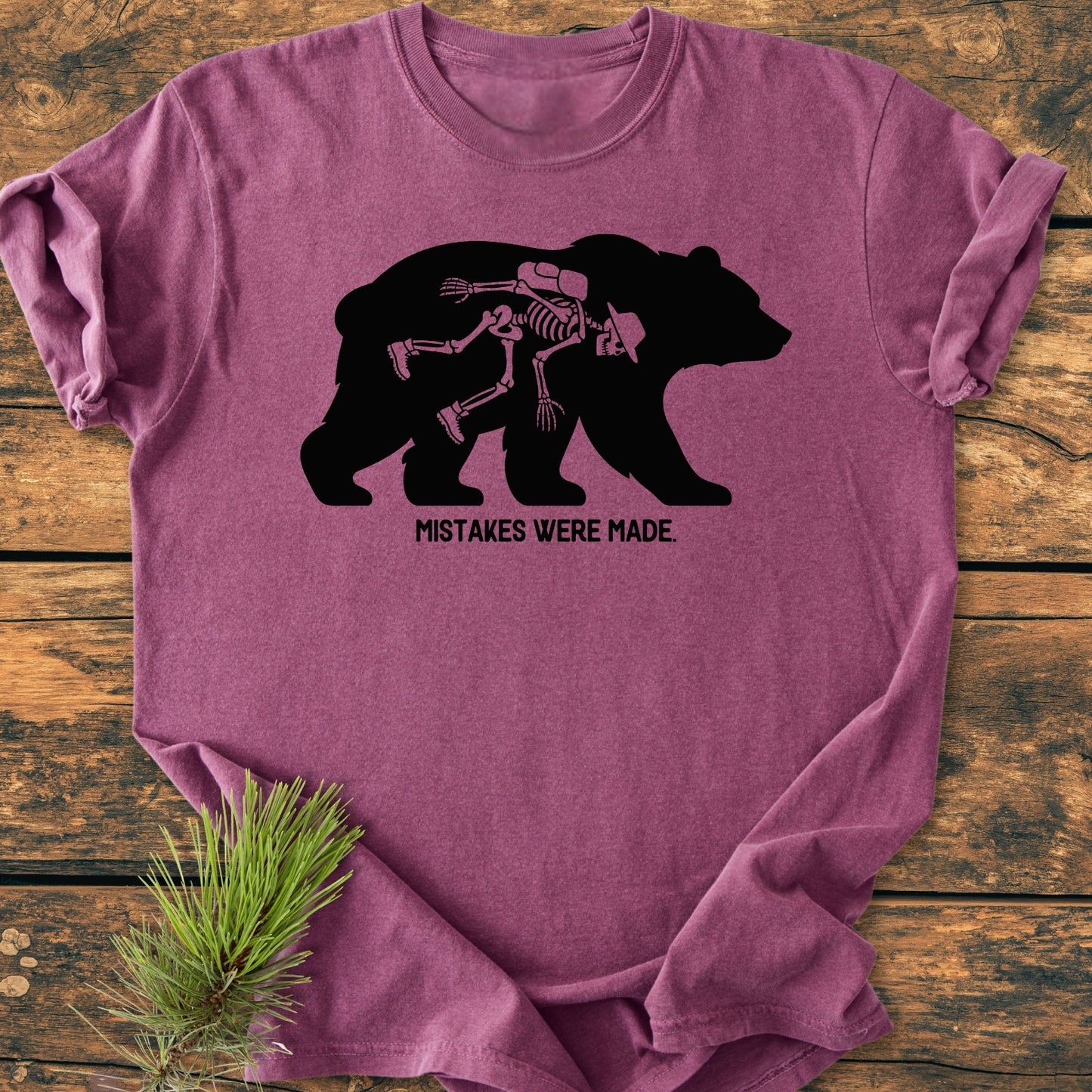 Mistakes Were Made - Vintage Wash Tee featuring a bear silhouette with a human skeleton inside, printed in black on a heather-purple shirt, laid on wood with a pine sprig accent.