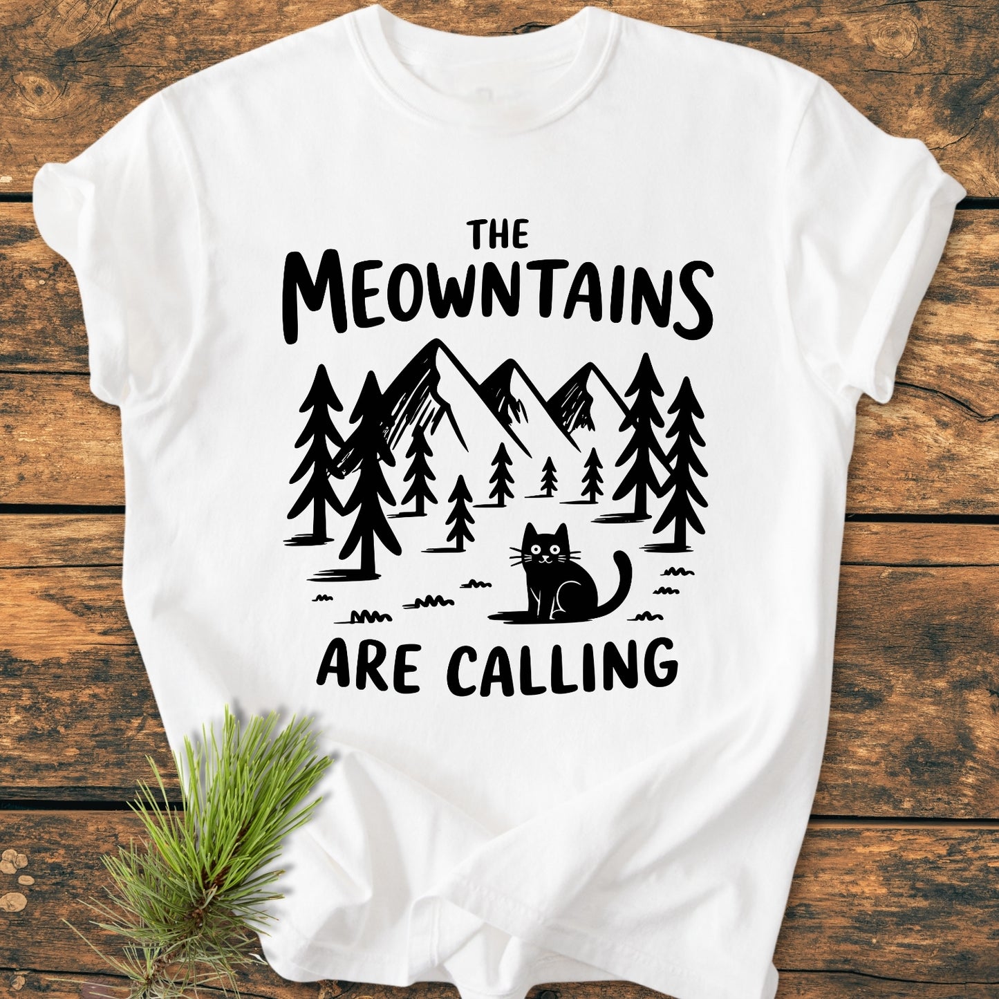 The Meowntains Are Calling - Vintage Wash Tee features a black mountain-and-pine scene with a cat on a white T-shirt, emphasizing comfort and durability with its relaxed fit and soft-washed fabric.