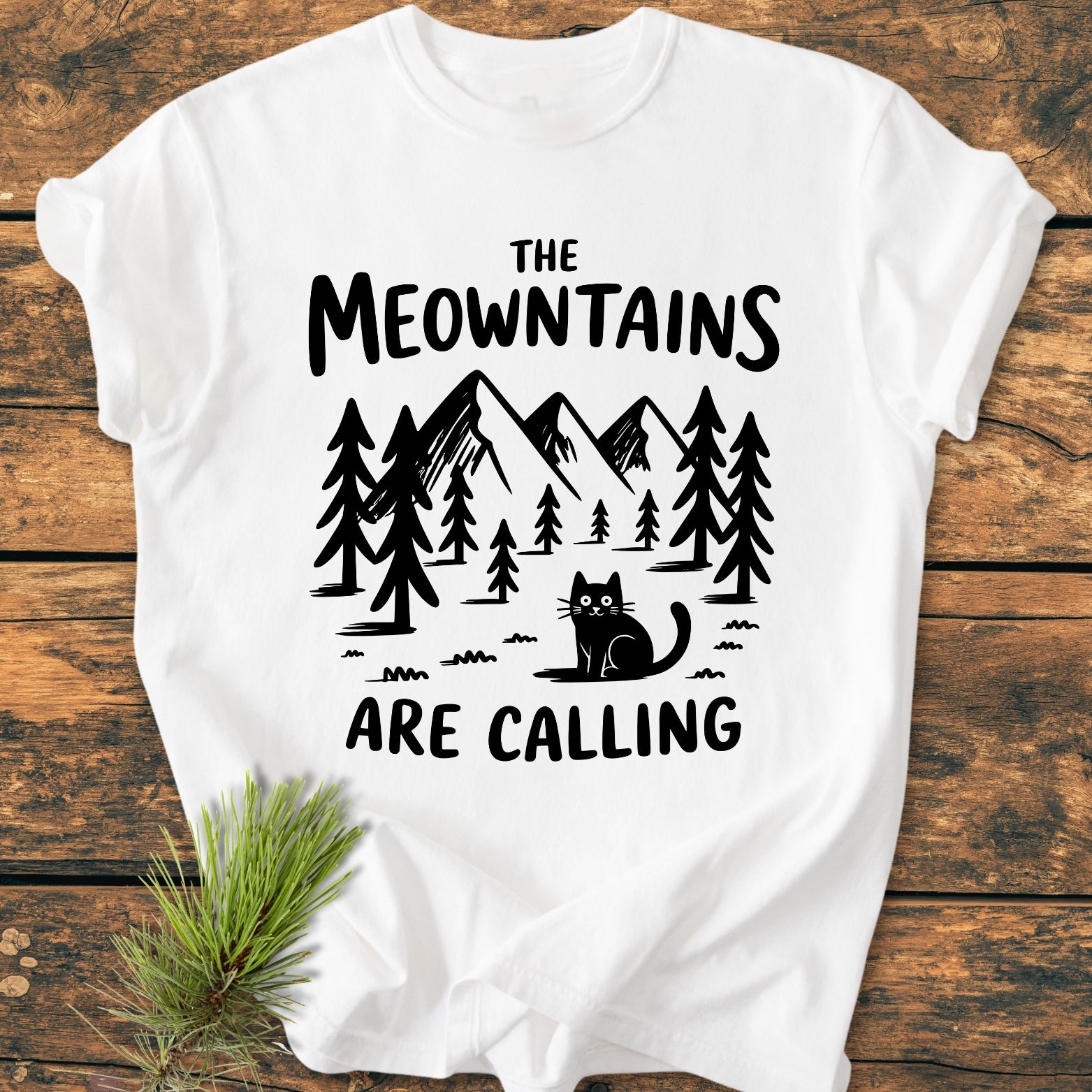 The Meowntains Are Calling - Vintage Wash Tee features a black mountain-and-pine scene with a cat on a white T-shirt, emphasizing comfort and durability with its relaxed fit and soft-washed fabric.
