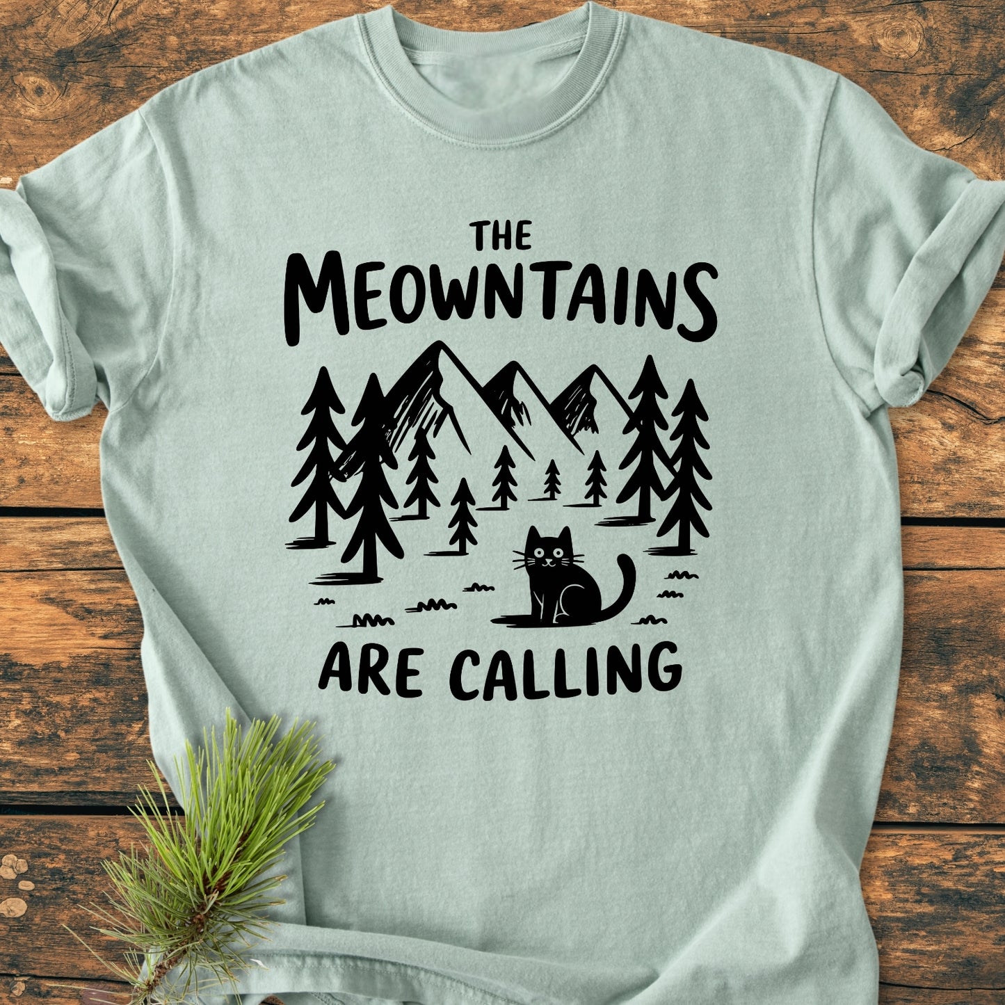 The Meowntains Are Calling - Vintage Wash Tee features a forested mountain graphic with a cat and slogan on light-green fabric, laid on wood with a pine sprig, highlighting its cozy, relaxed fit.