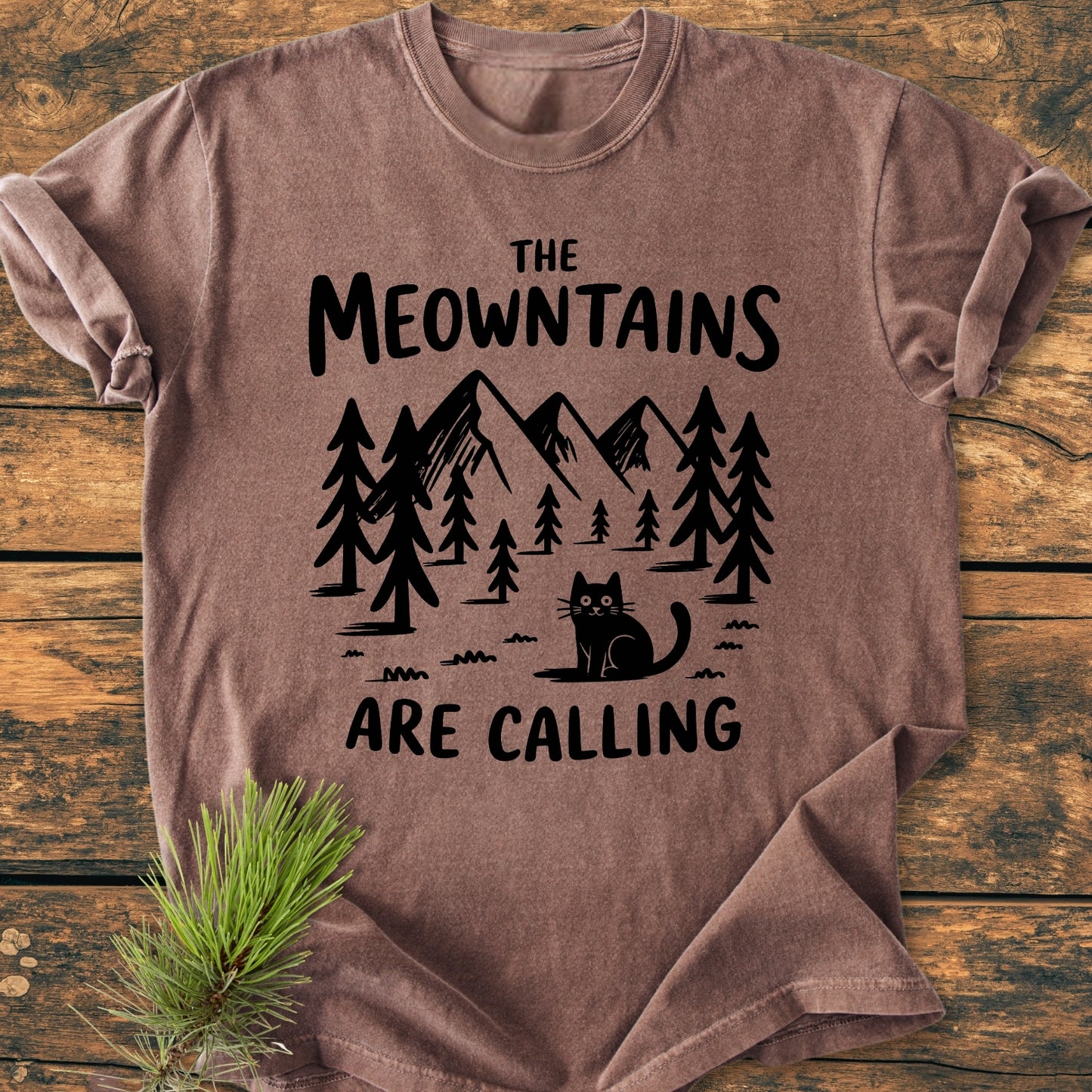 The Meowntains Are Calling - Vintage Wash Tee features a mountain-forest-cat graphic on a soft, relaxed-fit brown T-shirt, laid flat with a pine branch accenting the scene.