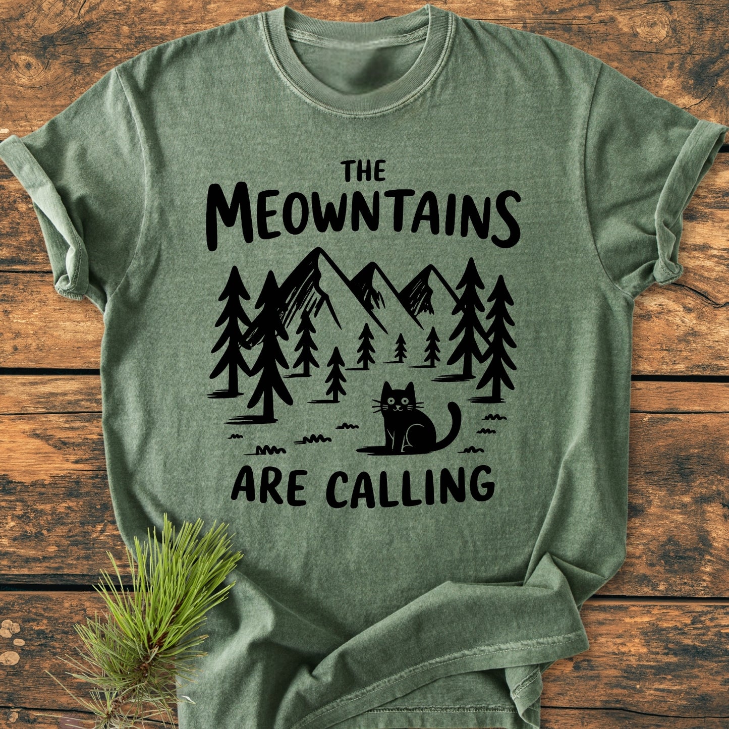 The Meowntains Are Calling - Vintage Wash Tee features a playful pine and mountain print with a cartoon cat. Laid flat on wood, it highlights Comfort Colors’ relaxed, durable garment-dyed fabric.