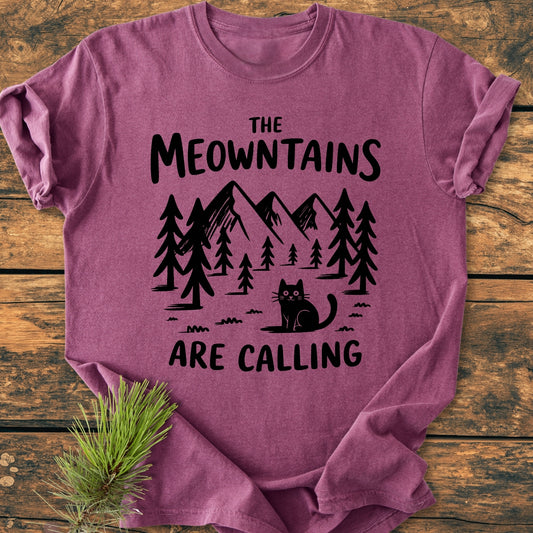 The Meowntains Are Calling - Vintage Wash Tee features a black graphic of pine trees, mountain peaks, and a cat, with a pun slogan, laid on a wooden surface with a pine sprig.