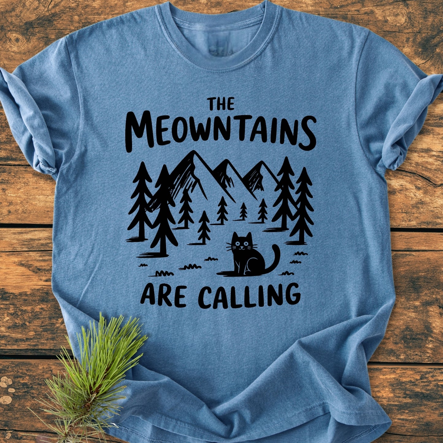 The Meowntains Are Calling - Vintage Wash Tee displays a stylized mountain-and-forest scene with a cat, printed on a heather-blue T-shirt, laid on a wooden surface with pine nearby.