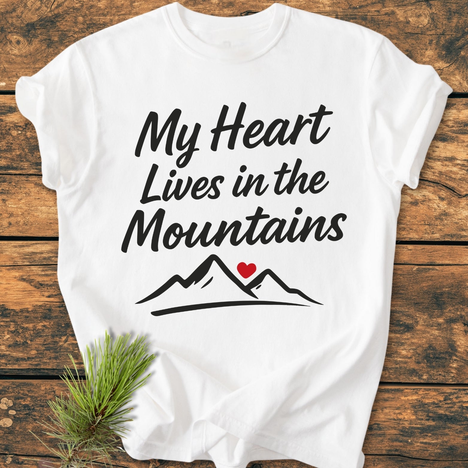 My Heart Lives In The Mountains - Vintage Wash Tee on wooden surface, featuring bold script with mountain outline and red heart. Includes a sprig of pine next to the garment-dyed, relaxed fit T-shirt.