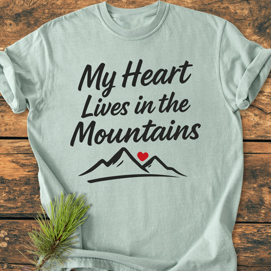 My Heart Lives In The Mountains - Vintage Wash Tee features a black script with mountain peaks and a red heart, laid on wood with a pine sprig, emphasizing comfort and durability.
