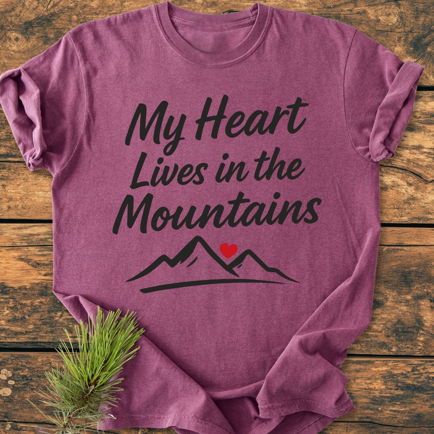 My Heart Lives In The Mountains - Vintage Wash Tee features a heathered purple design with mountain peaks and heart graphic, laid on wood, complemented by pine sprig, emphasizing its outdoorsy theme.