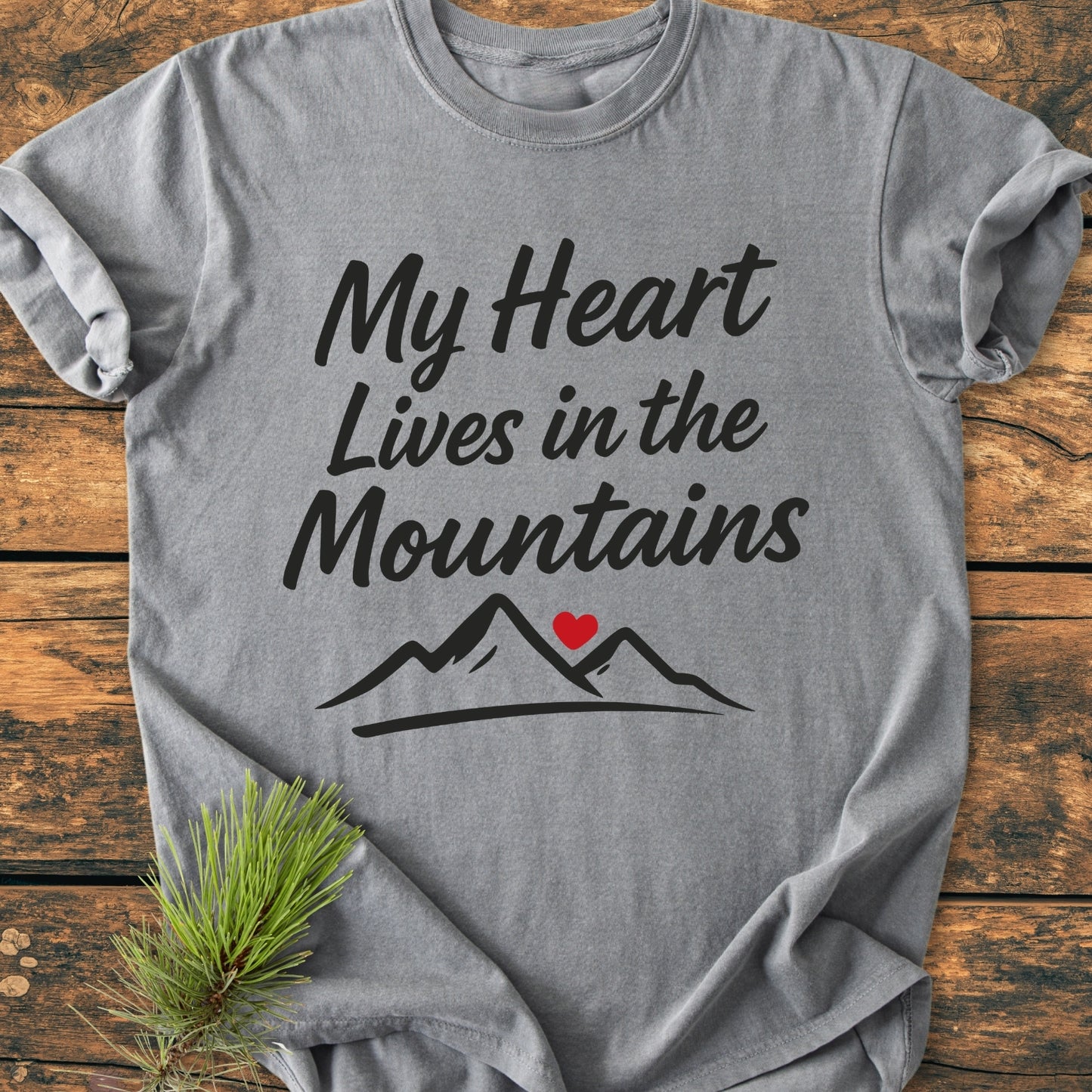 My Heart Lives In The Mountains - Vintage Wash Tee on wooden surface, featuring hand-lettered text and mountain graphic with heart. Styled with pine branch, showcasing its relaxed fit and garment-dyed fabric.