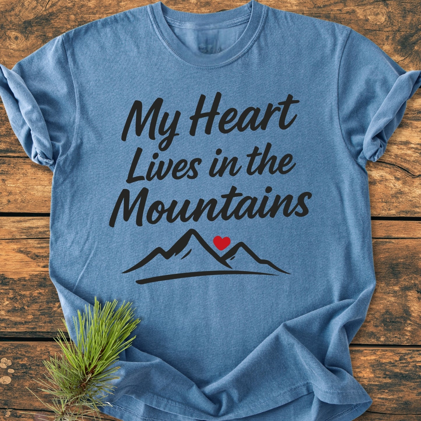 My Heart Lives In The Mountains - Vintage Wash Tee features a heather blue crew-neck design with black script and mountain illustration, showcased on rustic wood with a pine needle accent.