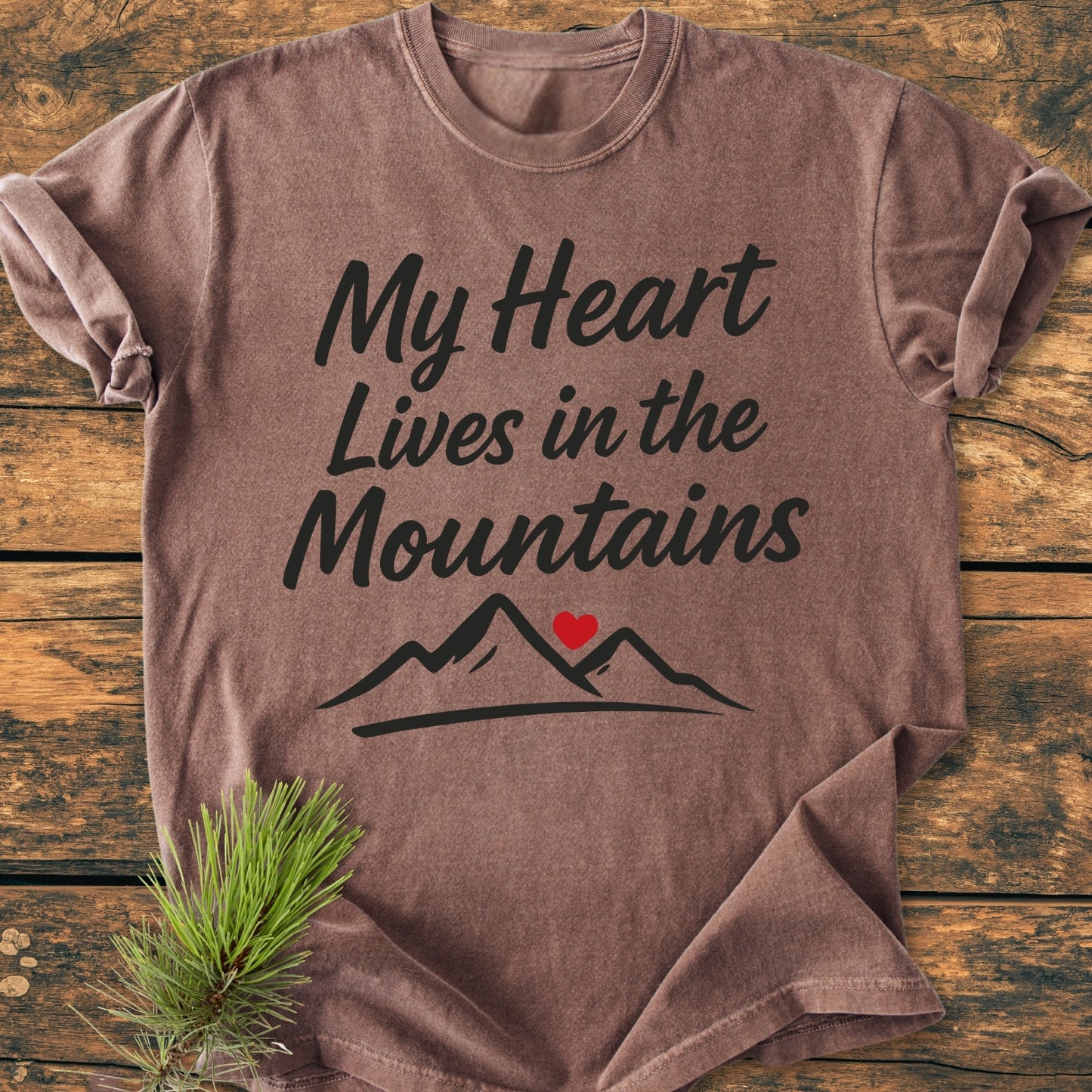 My Heart Lives In The Mountains Vintage Wash Tee on a wooden surface, featuring bold script and mountain design with a red heart. Pine sprig adds rustic, outdoorsy charm.