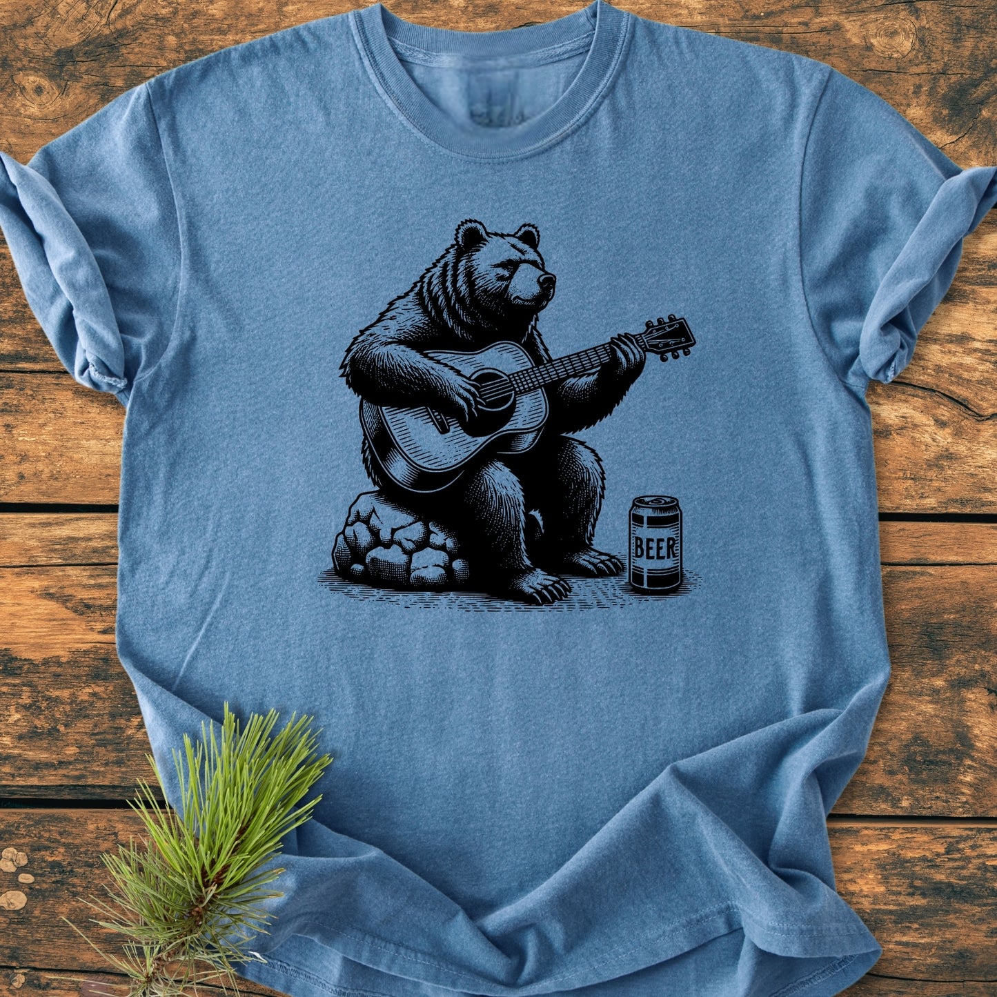 Bear Playing Guitar - Vintage Wash Tee features a black graphic of a bear with a guitar and beer can on a heather-blue, soft-washed T-shirt, laid flat on a wooden tabletop.