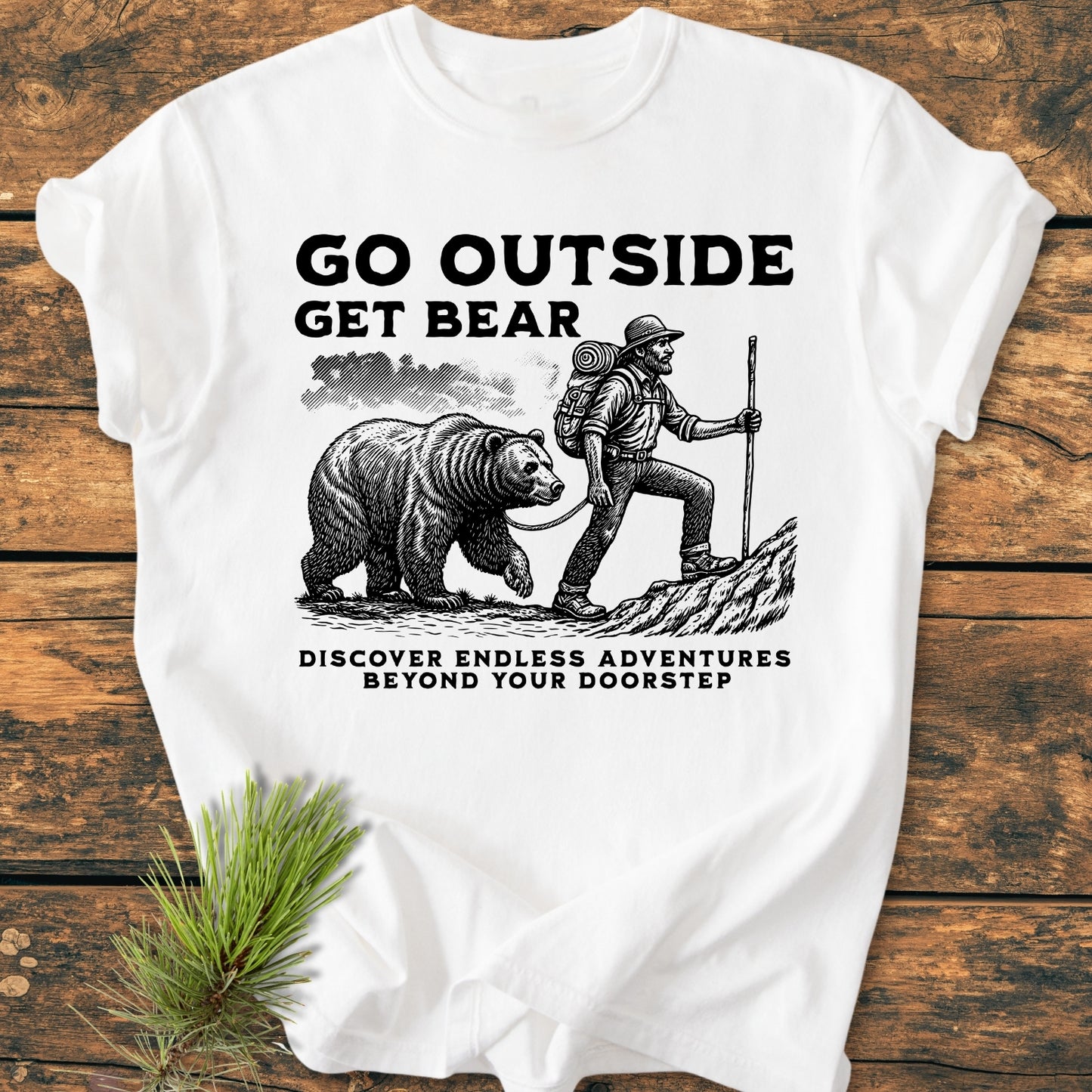 Go Outside Get Bear - Vintage Wash Tee features a rustic illustration of a hiker with a bear. Displayed on a wooden surface, it includes nature-themed pine sprigs for an adventurous touch.