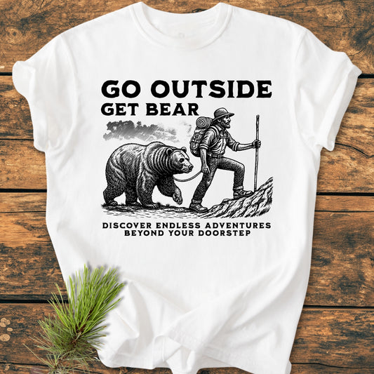 Go Outside Get Bear - Vintage Wash Tee features a rustic illustration of a hiker with a bear. Displayed on a wooden surface, it includes nature-themed pine sprigs for an adventurous touch.