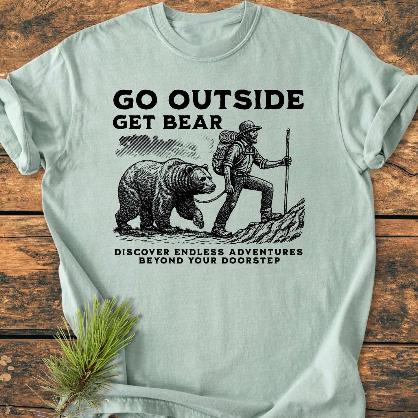 Go Outside Get Bear - Vintage Wash Tee features a backpacker illustration with a bear on a leash, printed on a soft, relaxed-fit pale-green T-shirt, laid on a wooden surface with pine sprig.