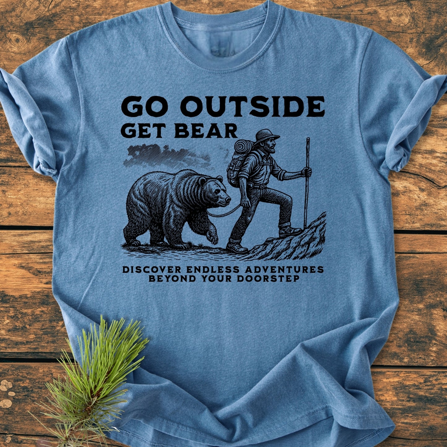 Go Outside Get Bear - Vintage Wash Tee displays a whimsical hiker with a bear illustration, surrounded by nature-inspired text. Laid on wooden surface, highlighting its cozy, relaxed fit and durable craftsmanship.