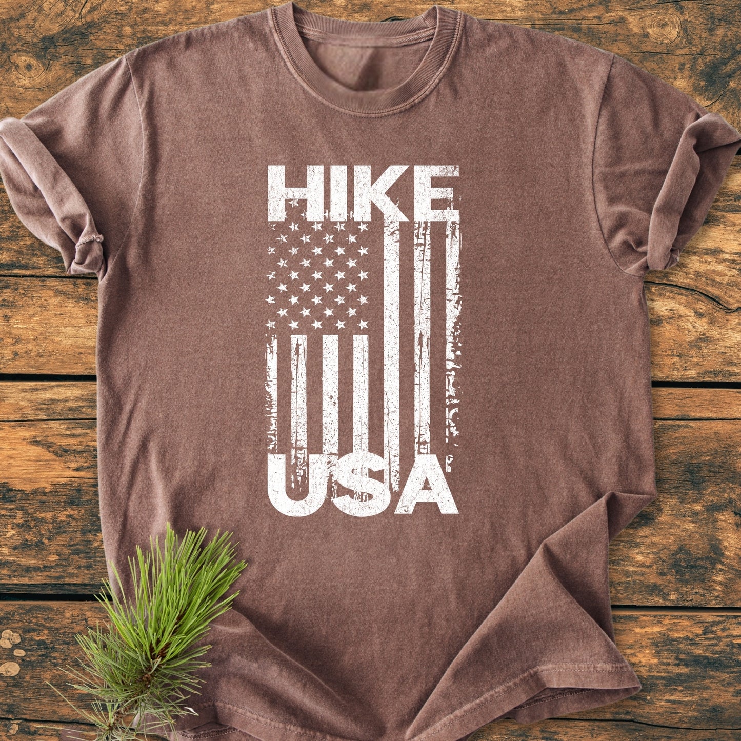 Hike USA - Vintage Wash Tee features a distressed white American flag design on a brown cotton T-shirt, displayed on a wooden plank background, with a pine bough accentuating its rustic charm.