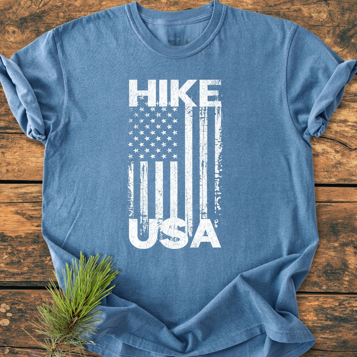 Hike USA - Vintage Wash Tee in heather-blue features a distressed vertical American flag graphic. Laid flat on wood, it highlights its relaxed fit and soft, garment-dyed cotton fabric.