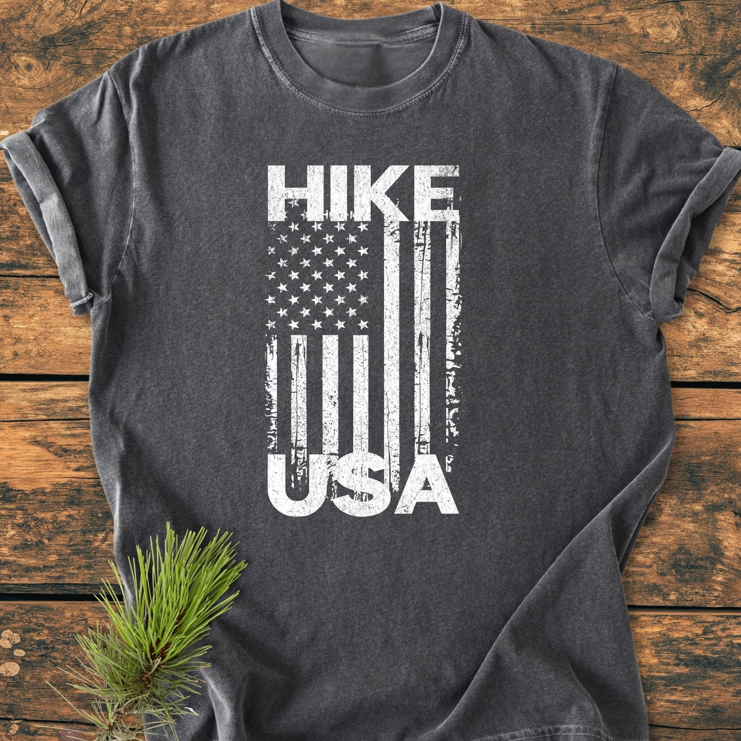 Hike USA - Vintage Wash Tee: A dark gray T-shirt with a worn white vertical American flag graphic, HIKE above, USA in stripes, laid flat on wood, complemented by a small evergreen sprig.