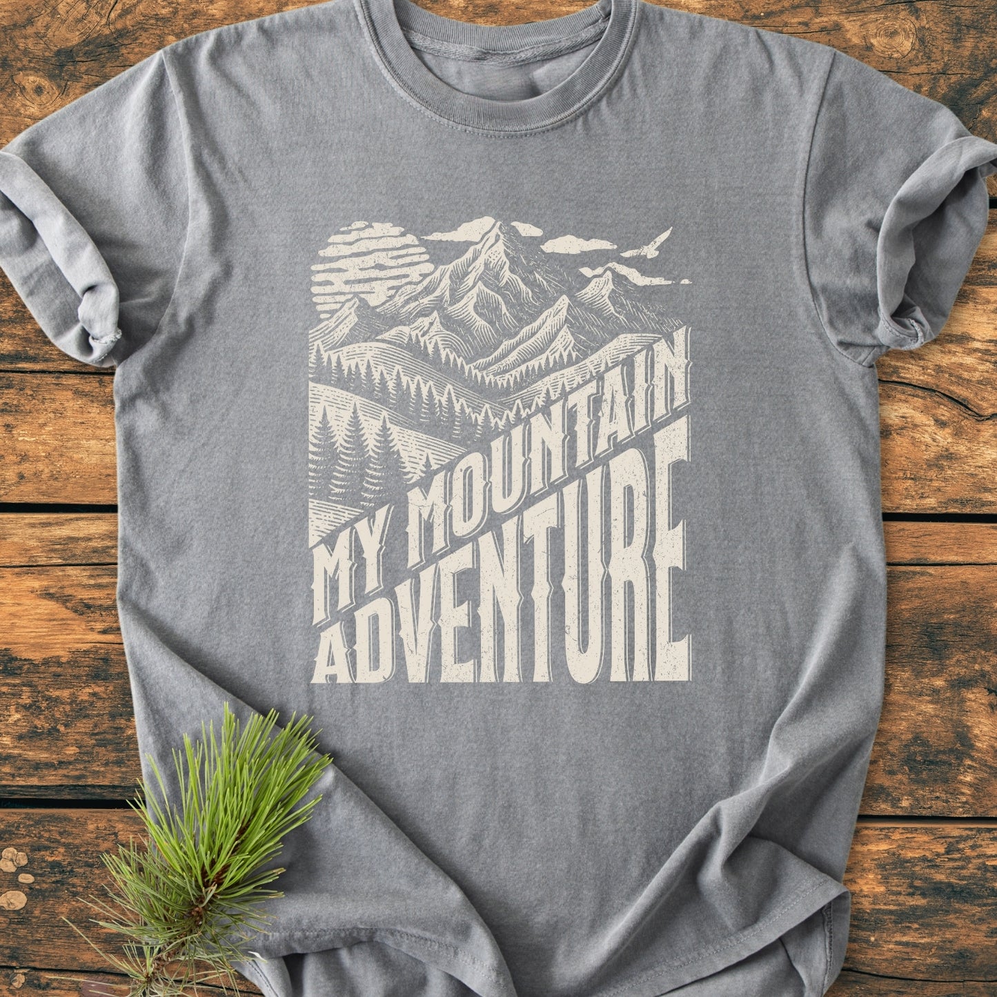 My Mountain Adventure - Vintage Wash Tee features a cream mountain graphic with sun and bird on a heather-gray T-shirt, laid on wood, accented by a pine sprig.