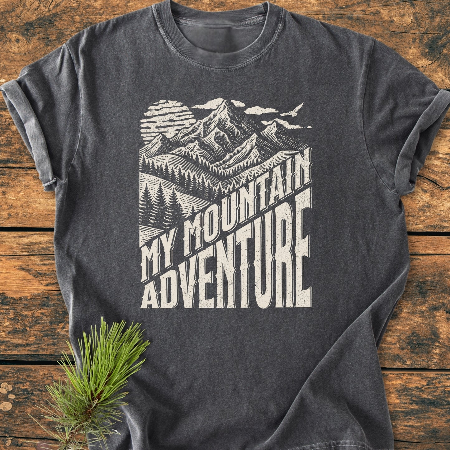 Charcoal-gray My Mountain Adventure - Vintage Wash Tee with a vintage-style graphic of mountains, sun, pine trees, and bold text, laid flat on a wooden surface, showcasing rustic, outdoorsy charm.