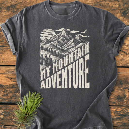 Charcoal-gray My Mountain Adventure - Vintage Wash Tee with a vintage-style graphic of mountains, sun, pine trees, and bold text, laid flat on a wooden surface, showcasing rustic, outdoorsy charm.