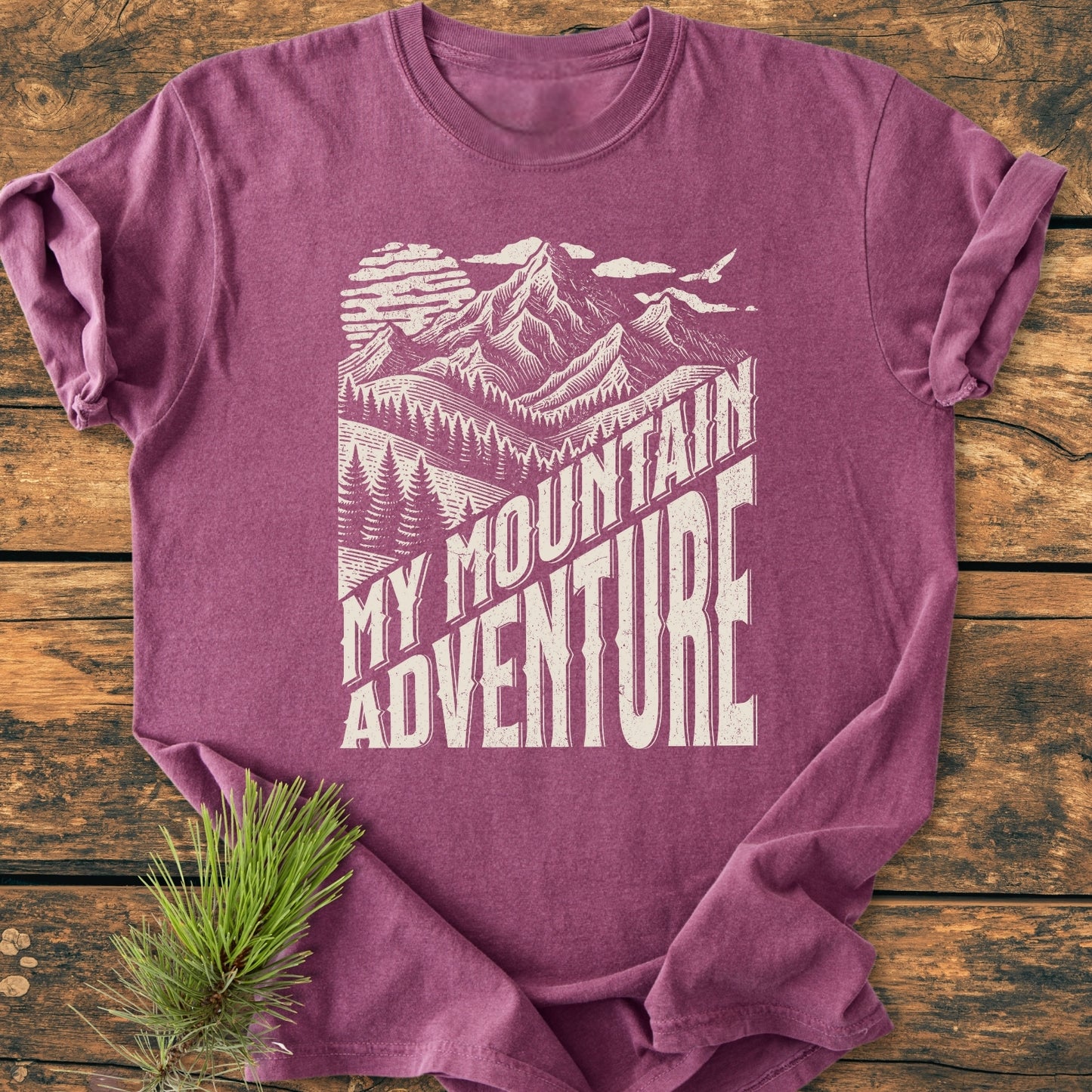My Mountain Adventure - Vintage Wash Tee features a heather-purple shade with a printed mountain scene, laid on wood, highlighting its cozy, relaxed fit and durable construction with double-needle stitching.